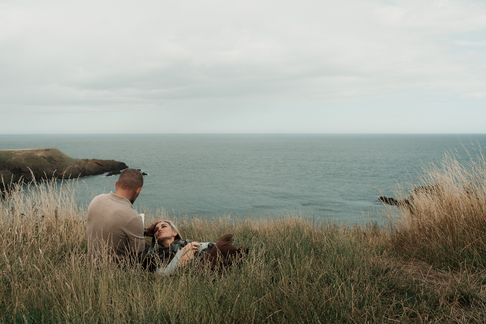 A couple lying together in long grass on a clifftop above the sea